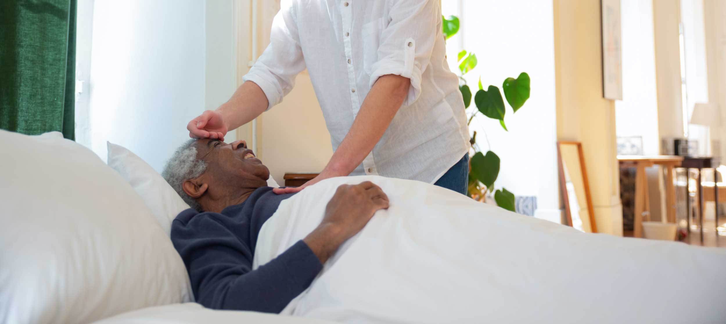 A person gently touches the forehead of an elderly man lying in bed. The man is covered with a white blanket and appears to be resting comfortably. Sunlight streams into the room, illuminating green plants and creating a calm, peaceful atmosphere.
