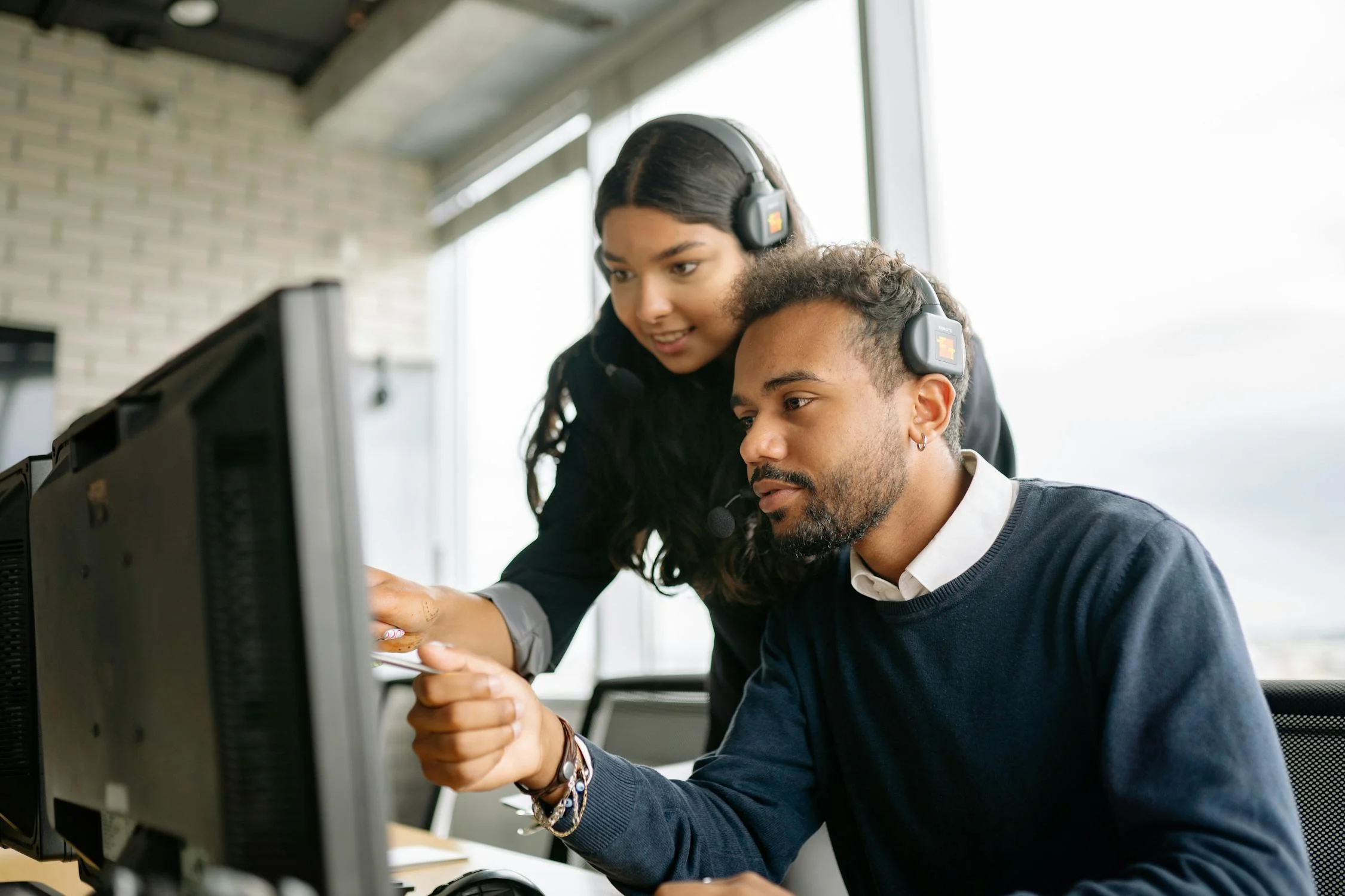 Customer service workers with headsets assist eachother with performing computer tasks.