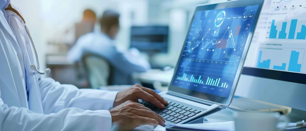 A person wearing a white medical coat uses a laptop showing various charts and graphs on the screen. Other individuals work at desks in the background, with monitors displaying similar data. The setting appears to be a modern office or healthcare facility.