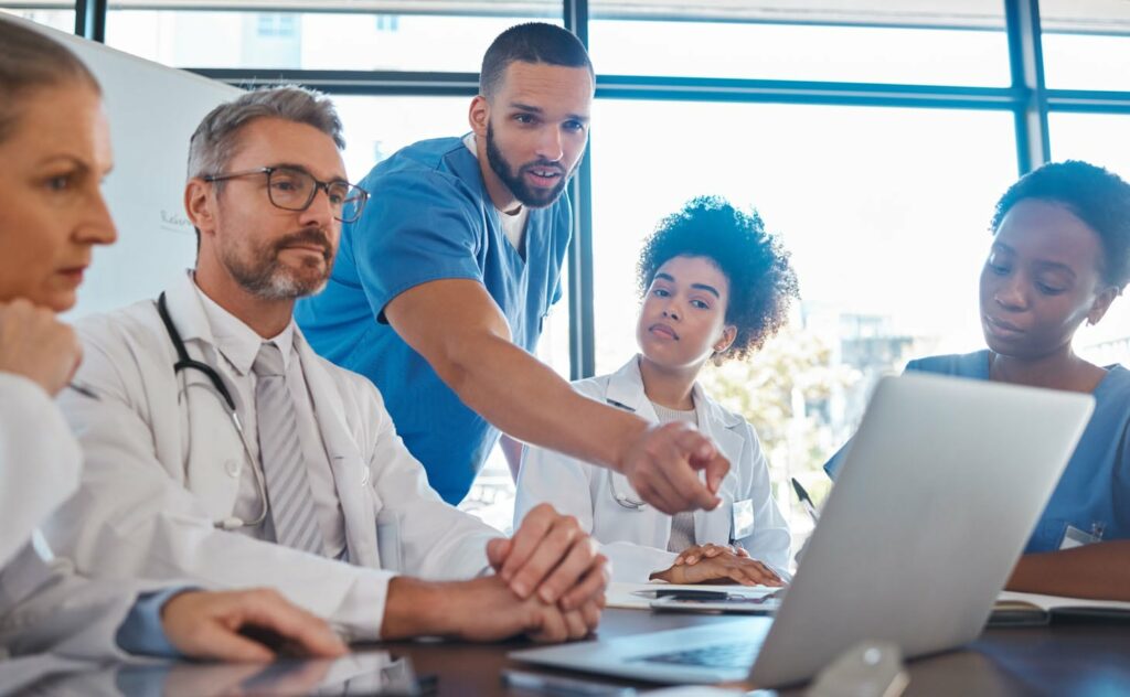 A group of five medical professionals gathered around a table in a meeting. One person, in scrubs, stands and points at a laptop screen while explaining something to the others. The rest, in lab coats or scrubs, listen attentively.