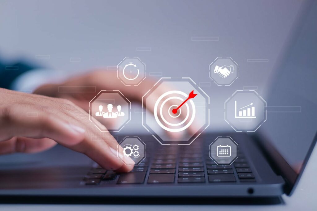 Close-up of hands typing on a laptop keyboard. Transparent icons in front of the laptop include a target with an arrow, a handshake, a clock, a bar graph, a group of people, gears, and a calendar, representing business and productivity concepts.