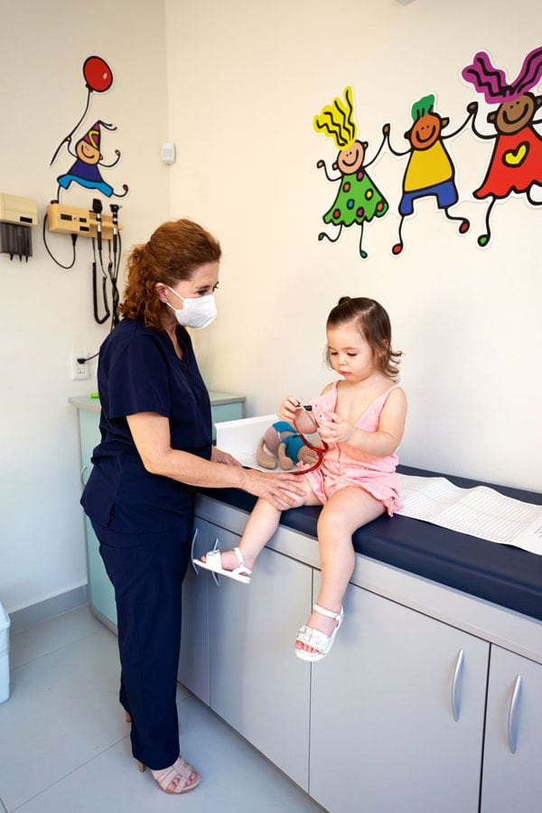 A healthcare professional in navy blue scrubs and a face mask is examining a young child sitting on an examination table in a medical office. The child is wearing a light pink outfit and is holding a toy. The wall behind them is decorated with colorful drawings of happy children.