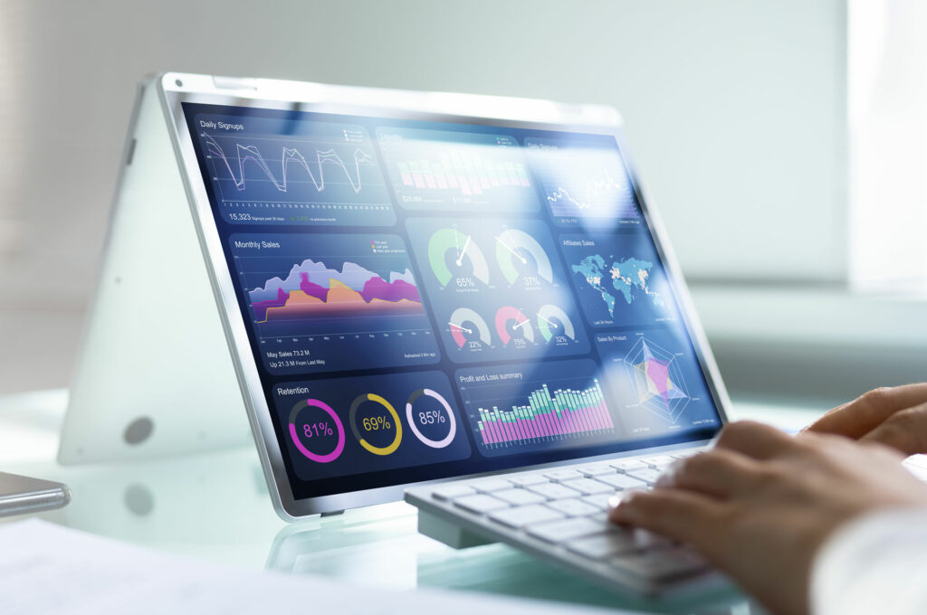 A person works on a tablet displaying various data visualizations and charts, including line graphs, pie charts, bar graphs, and a world map. A wireless keyboard is positioned in front of the tablet on a white desk.