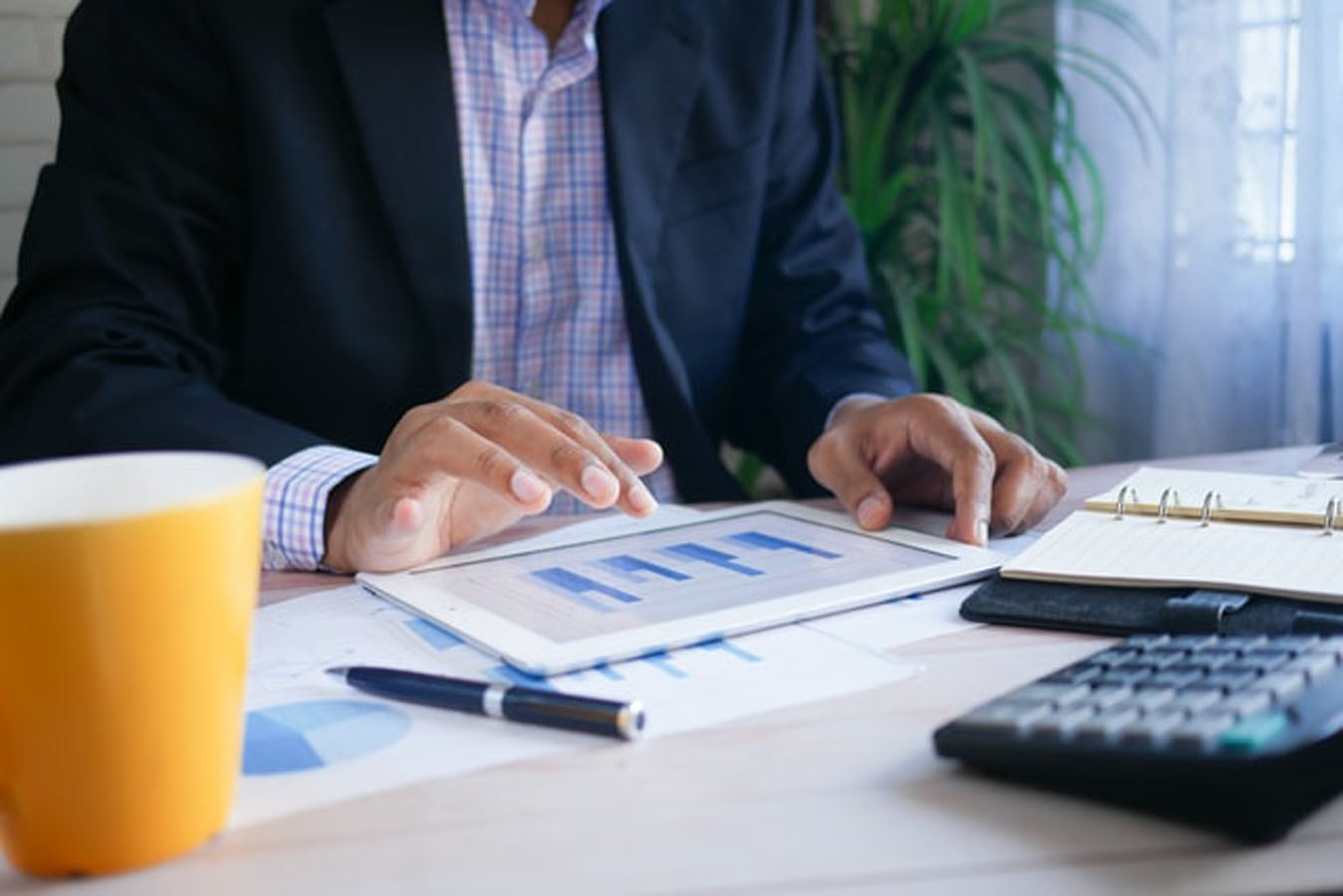 A person in a business suit uses a tablet displaying a bar graph at a desk. The desk has a yellow coffee mug, pen, calculator, and paperwork with charts. A potted plant is in the background.